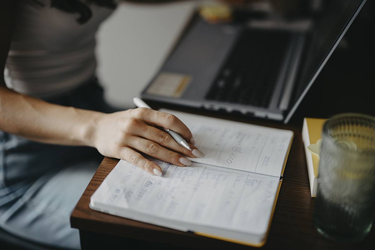 Ruka píše poznámky do sešitu vedle otevřeného notebooku